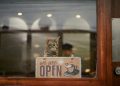 A cute tabby cat sits behind a cafe window, peering over an 'Open' sign.